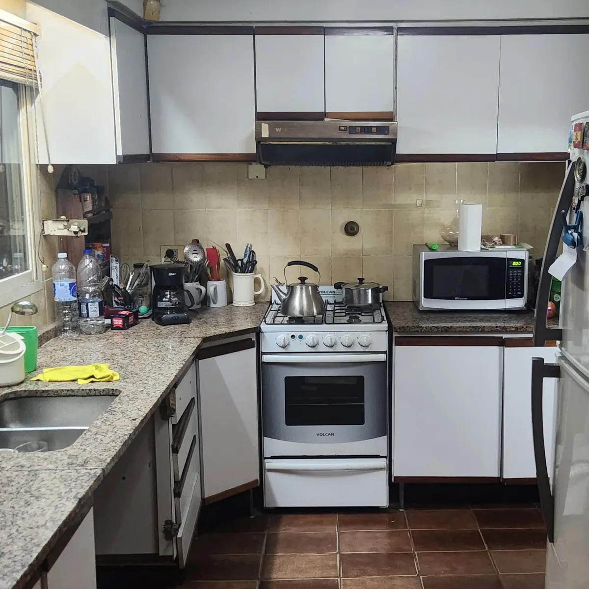 La cocina antes de la obra: mesada de granito en U, alacenas blancas hasta el techo, azulejo beige, cocina a gas con horno y la heladera contra el paso