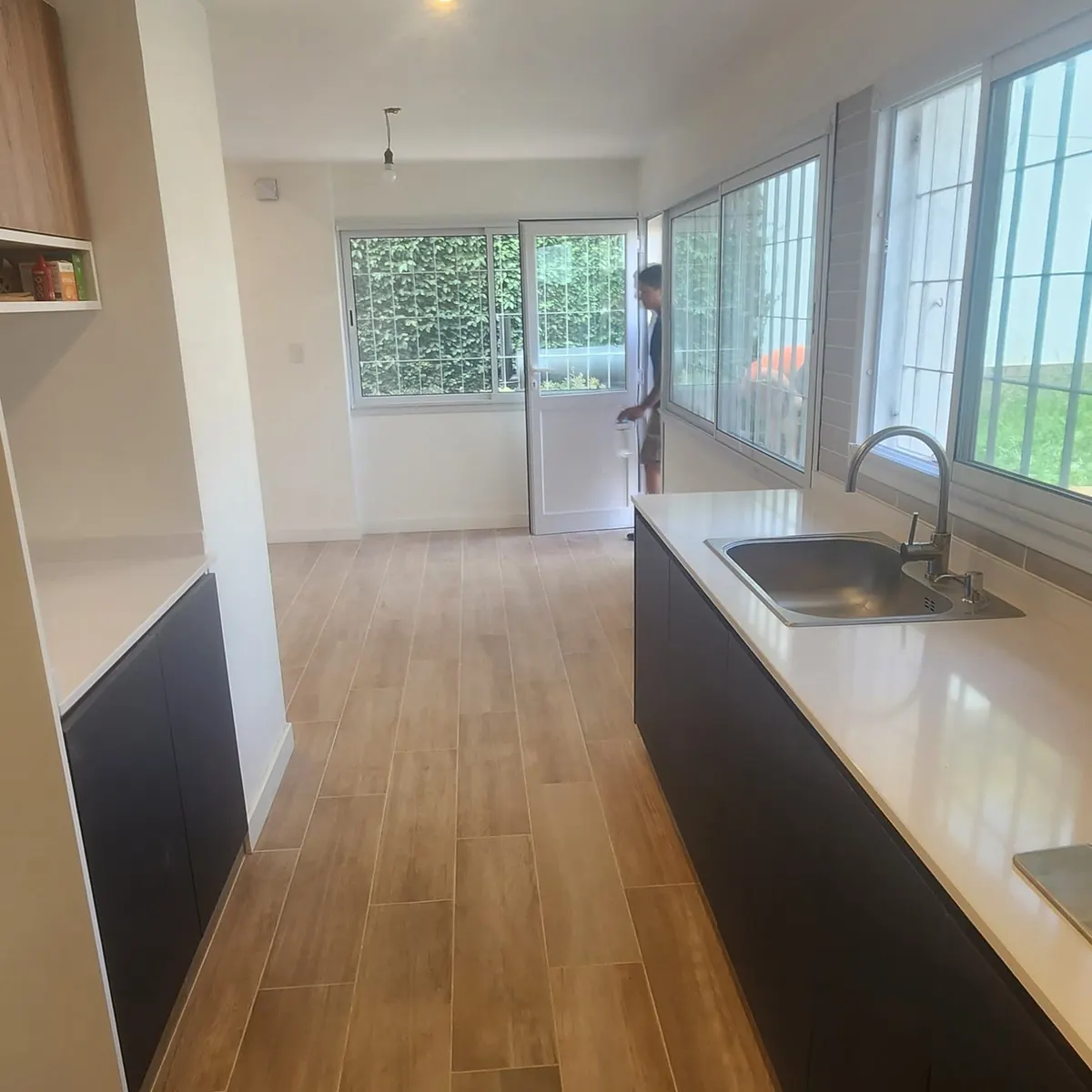 La cocina nueva, alargada: mesada blanca con bacha de acero de un lado, muebles bajos oscuros y, al fondo, el comedor con el ventanal en esquina y la puerta al jardín