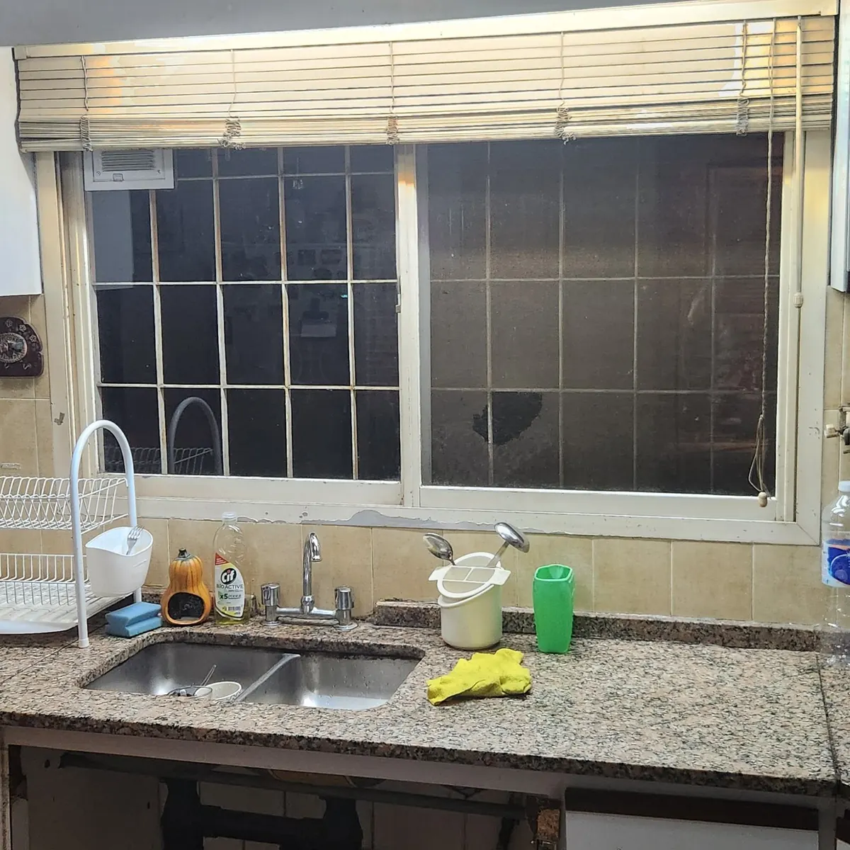 La mesada vieja de granito con la bacha doble de acero bajo una ventana con reja y cortina de enrollar