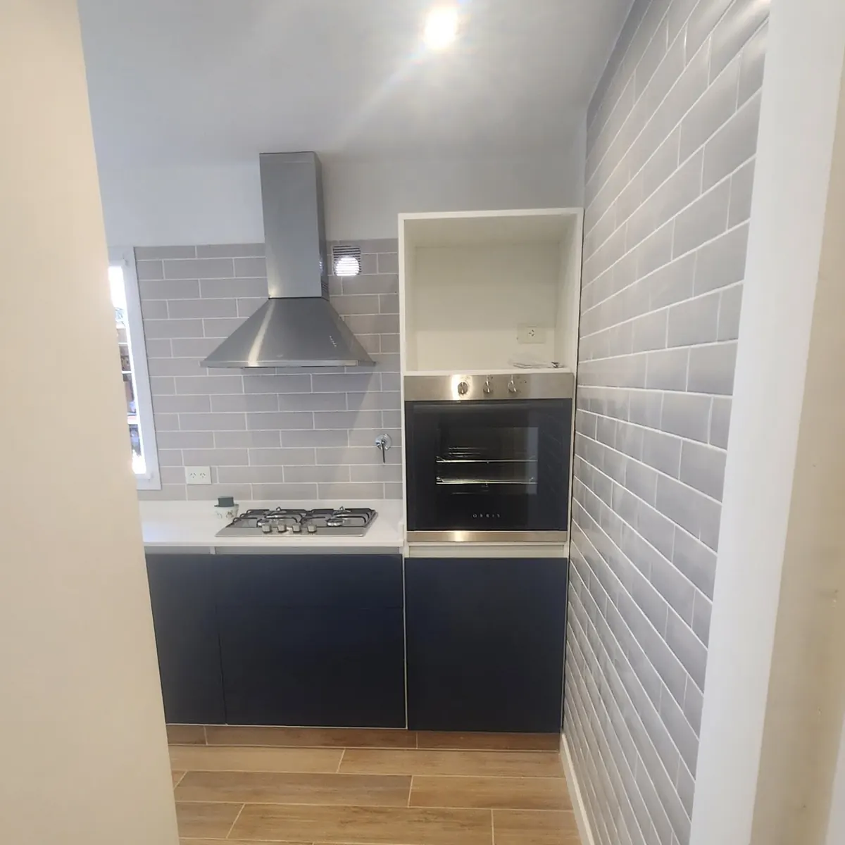 El sector de cocción de la cocina nueva: azulejo tipo subway gris, campana de acero inoxidable, anafe sobre un mueble bajo oscuro y horno empotrado en columna