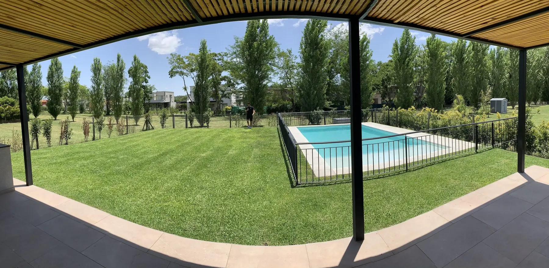 Panorámica desde la galería: el alero de listones de madera sobre las columnas negras y el parque de lado a lado