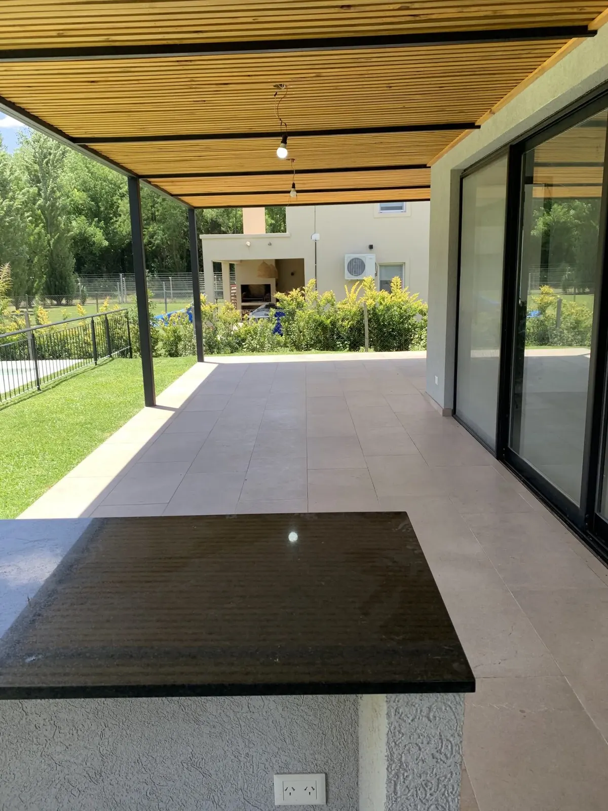 La galería terminada: mesada de granito negro en primer plano, piso de porcelanato gris de gran formato y cielorraso de listones de madera sobre columnas de acero negro