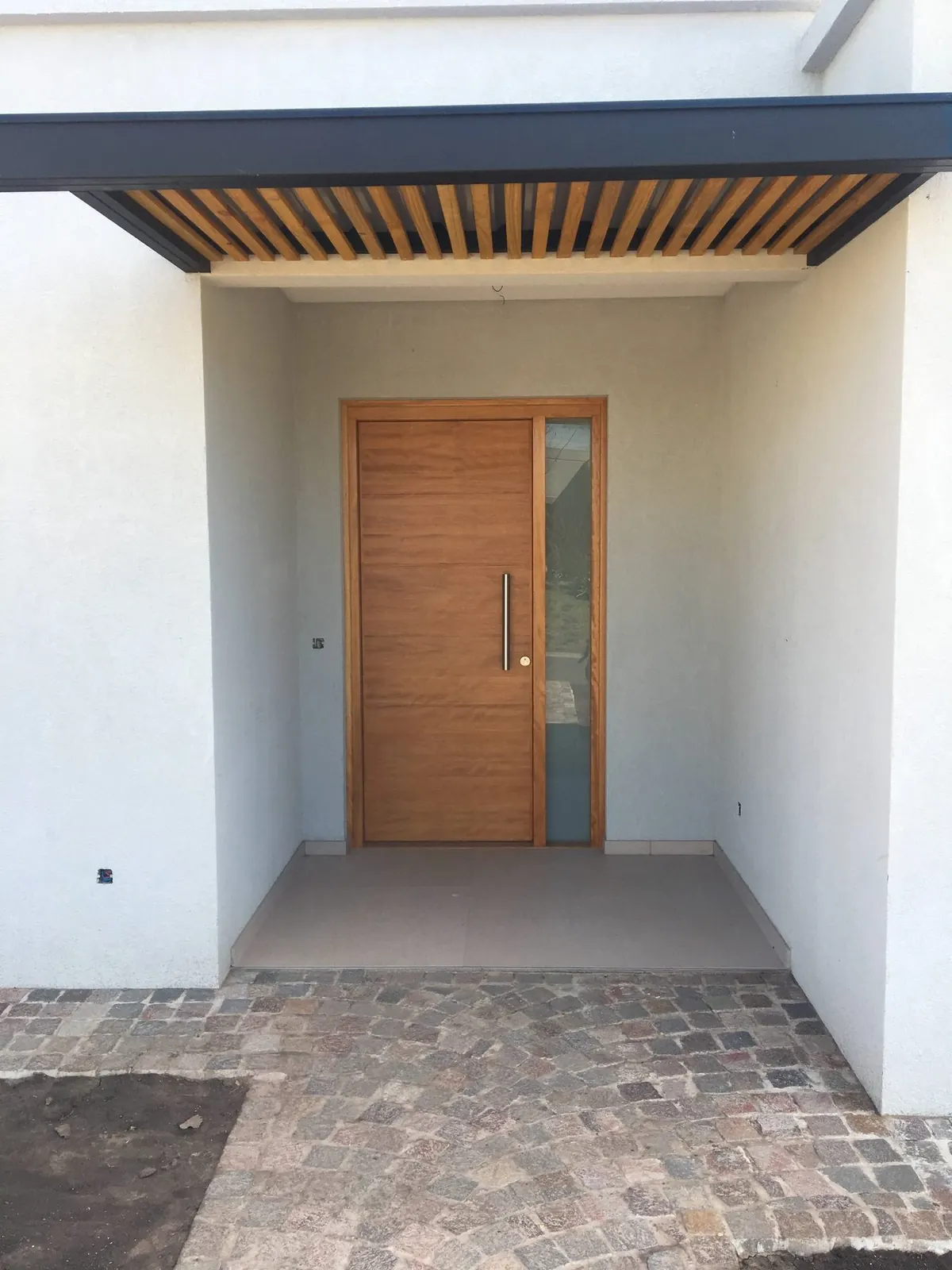 El acceso: puerta de madera maciza con paño fijo lateral, bajo el alero de listones de madera